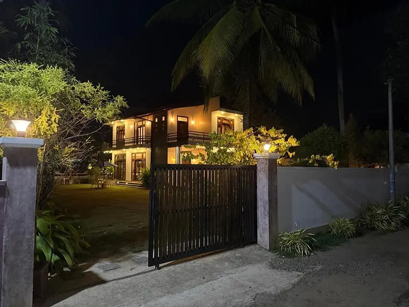 The villa gate and driveway at night, the house lit behind the palms