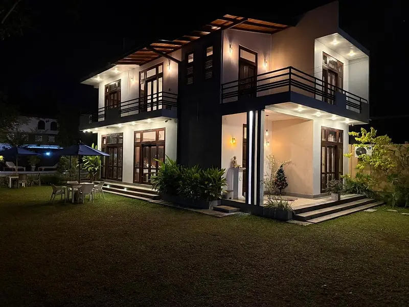The white two-storey villa at night, warm lights on the balconies and the garden in front