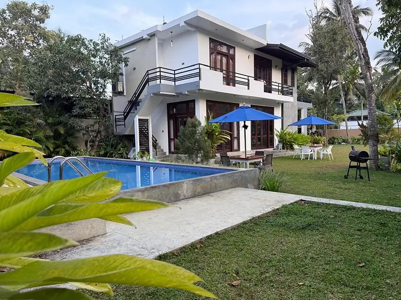 The villa seen across the blue pool and lawn on a sunny day, with blue umbrellas in the garden